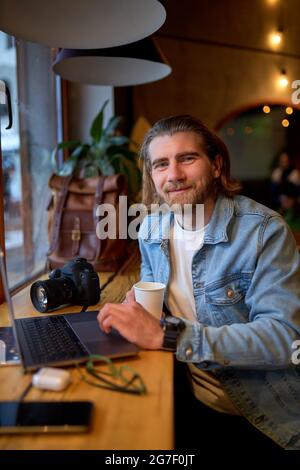 Portrait of positive bearded guy looking at camera while working on laptop in cafe, looking pleasant and nice, wearing denim jacket, casual outfit. ca Stock Photo