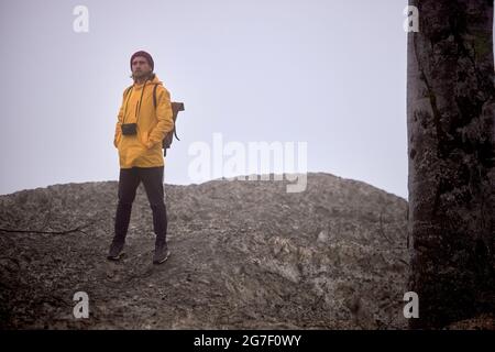 Handsome traveler Male with camera walking down the mountain, at foggy weather, alone , in yellow coat, stands looking at side, in wild nature, advent Stock Photo