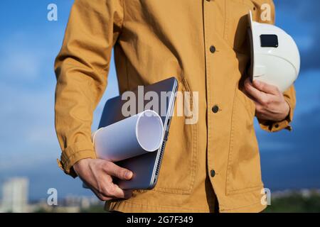 Supervisor, foreman, manager or building contractor in construction industry concept. Male in orange jacket standing and holding hard hat, laptop and blueprints. Engineer or worker on site Stock Photo