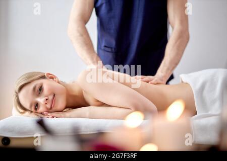 Charming Woman Lying On Belly Getting Spa Massage On Back By Male Masseur, Blonde Lady Of Caucasian Appearance Looking At Camera, Relaxed, Leisure Tim Stock Photo