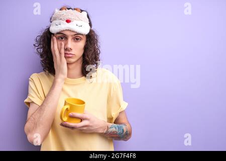 Student has no enough sleep, want to go in bed in the morning, dressed casually with eyemask for sleeping, holding yellow mug in hands Stock Photo