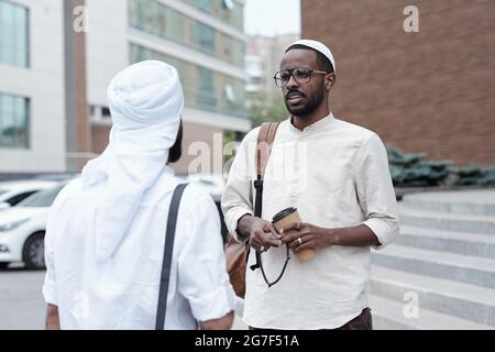 Confident young Muslim man in eyeglasses standing on street and drinking coffee with business partner Stock Photo