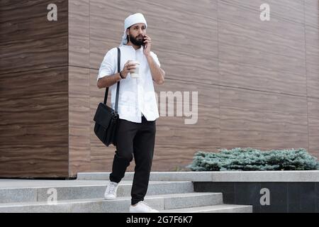Pensive young bearded Muslim businessman in ghutrah moving down stairs and talking by phone Stock Photo
