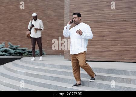 Serious young Middle Eatern man in white shirt moving down stairs and talking by phone Stock Photo