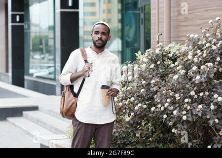 Pensive young African American man in kufi cap carrying satchel and drinking coffee on move Stock Photo