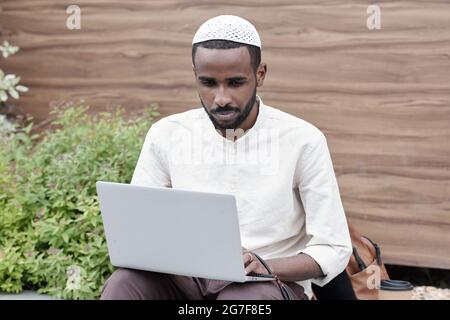 Content young Black man in muslim cap sitting outdoors and using laptop while working with social media Stock Photo