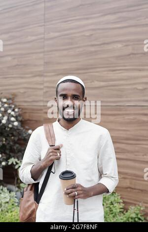 Portrait of smiling handsome young Islamic student in kufi cap standing with satchel and takeout coffee cup Stock Photo