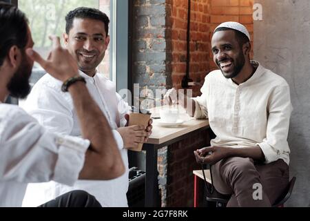 Group of jolly young Muslim friends drinking coffee and laughing at joke of man while gathering In cafe Stock Photo