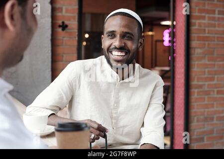Jolly handsome African American man in muslim cap holding rosary beads and chatting with friend in cafe Stock Photo