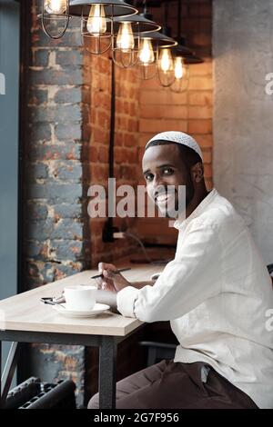 Portrait of smiling handsome bearded Black guy in kufi hat sitting with pen at table in loft cafe Stock Photo