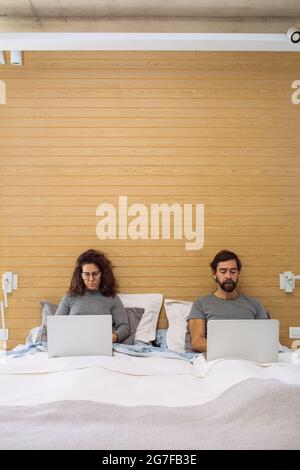 Couple in a double bed king size each with their notebook working watching each of their sreens. Concept: home office working from bed Stock Photo