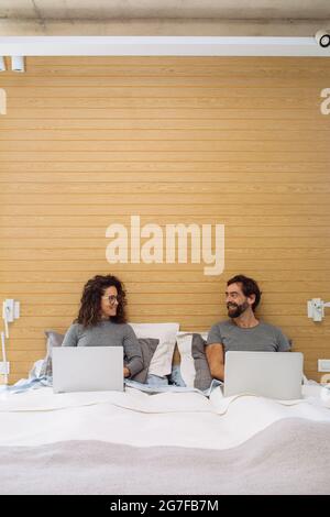 Couple in a double bed king size each with their notebook working looking each other and smiling. Concept: home office Stock Photo