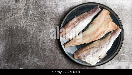 Raw Pollock (hake) fish fillet on a round plate on a dark gray background. Top view, flat lay Stock Photo