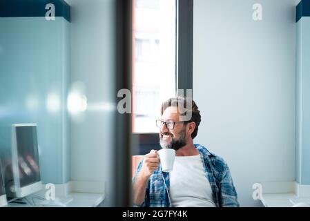 Portrait of happy man looking outside the window and smile drinking a coffee at home ot office - adult caucasian male with beard and glasses in job br Stock Photo