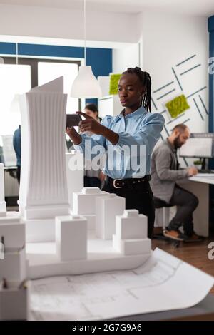 Woman of african american ethnicity working as architect with building plans maquette looking at smartphone for creative design. Worker viewing construction project for industry Stock Photo