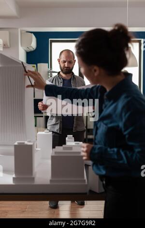 Architecture workers checking blueprints with maquette building model design plan. Caucasian workmates pointing at construction project layout for urban development business Stock Photo