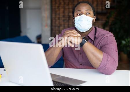 Pensive African-American guy wearing medical mask looks away sitting at the desk in front of laptop, a black male entrepreneur worried about crisis in business due to quarantine Stock Photo