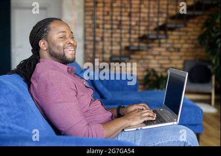 Inspired African-American man using laptop sitting on the couch at home, cheerful biracial guy with locks hair looks away dreamly, male writer journalist writing an article, waiting for inspiration Stock Photo