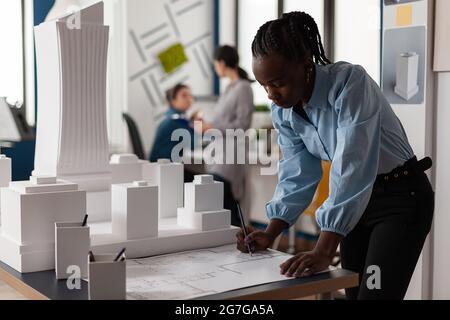 African american business woman at architectural office working on blueprint at desk. Young constructor engineer designing building model maquette in professional environment Stock Photo