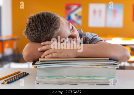 Tired caucasian schoolboy sitting at desk in classroom sleeping on a pile of schoolbooks Stock Photo