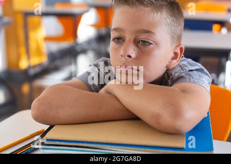 Tired looking caucasian schoolboy sitting at desk in classroom leaning on a pile of schoolbooks Stock Photo