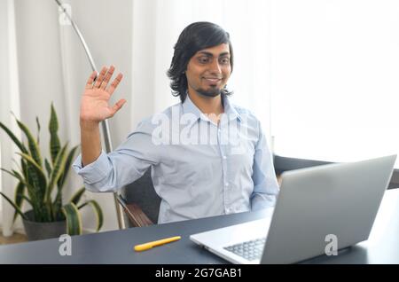 Eastern businessman looks at the webcam and waving, using laptop computer for video virtual meeting, greeting online interlocutor, sits in the contemporary office space Stock Photo