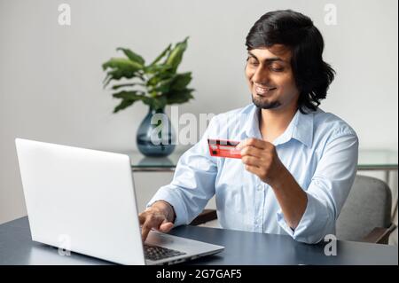 Cheerful and happy young indian man with credit card in hands making purchases online, using laptop computer for making online transaction, input data for paying online, e-banking concept Stock Photo