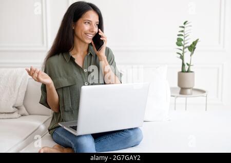Indian woman with laptop talking on mobile phone sitting on the comfortable couch at home. Mixed-race female freelancer talking on the smartphone. Student girl using laptop during phone conversation Stock Photo