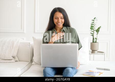 Cheerful indian woman using laptop for video call sitting on sofa at home, talking with friends or family on the distance, mixed-race female student glad to see online interlocutor, studying online Stock Photo