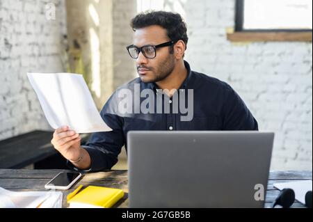 Executive mixed-race male office employee doing paperwork sitting at the desk with laptop in office, focused and serious hindu man in smart casual looking through sheets of documents, preparing report Stock Photo