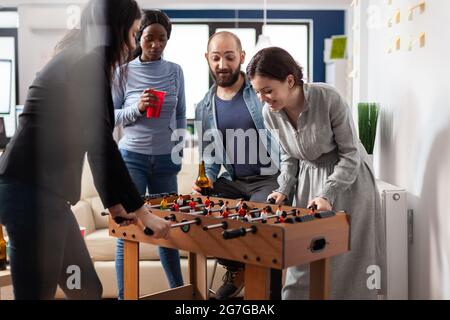 Multi ethnic group of people playing at foosball table after work partying and enjoying alcohol cups bottles of beer. Diverse team colleagues celebrating break from business activities Stock Photo