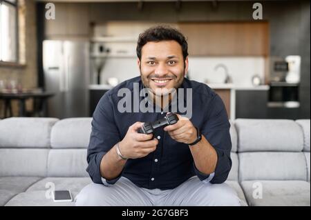 Handsome excited young indian man spends leisure time playing video game, hispanic guy with a joystick sitting on the couch, play on console at home, front view Stock Photo
