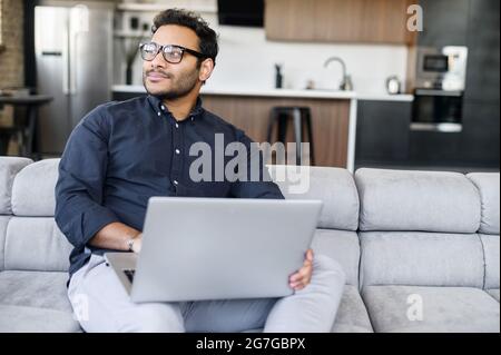 Handsome young indian freelancer man with beard in glasses sitting on the sofa and looking away, taking a break from working on laptop, dreaming about vacation or salary, enjoying working from home Stock Photo