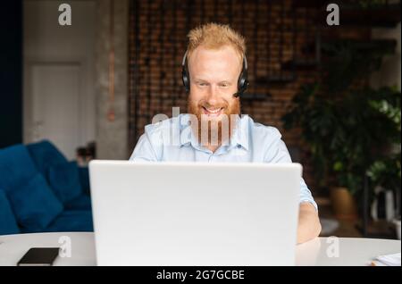 Portrait of cheerful red haired hipster guy in wireless headset using laptop computer sitting in contemporary loft style office space, man is making video call, talking online on distance, front view Stock Photo