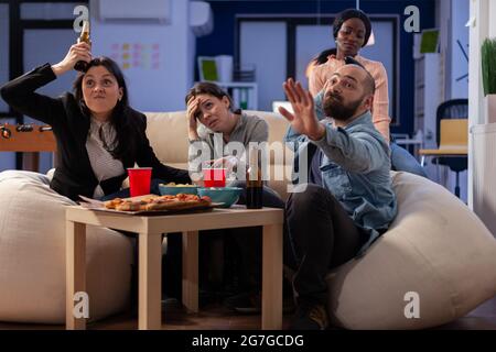 Team of multi ethnic friends losing tv game on console after work at office. Cheerful diverse group play with joystick while eating snacks pizza chips and drinking beer alcohol from cups Stock Photo