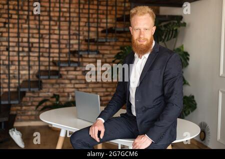 Successful red-haired businessman in formal suit leaned on the table in relaxed pose and looks at the camera, smiling male entrepreneur, ceo in loft style modern office Stock Photo