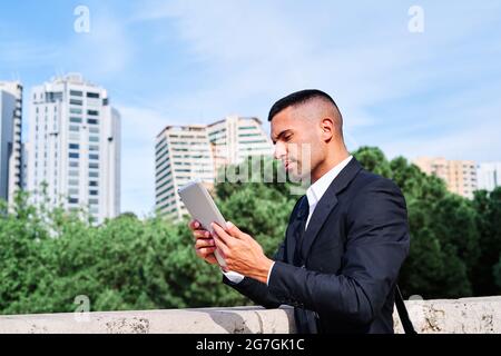 Side view of focused young Hispanic male executive manager in classy suit with bag reading information on tablet while standing near railing on urban Stock Photo