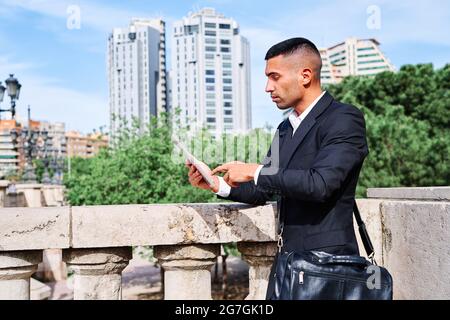 Side view of focused young Hispanic male executive manager in classy suit with bag reading information on tablet while standing near railing on urban Stock Photo