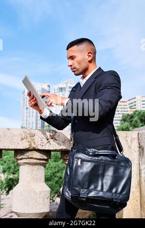 Side view of focused young Hispanic male executive manager in classy suit with bag reading information on tablet while standing near railing on urban Stock Photo