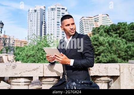 Side view of focused young Hispanic male executive manager in classy suit with bag reading information on tablet while standing near railing on urban Stock Photo