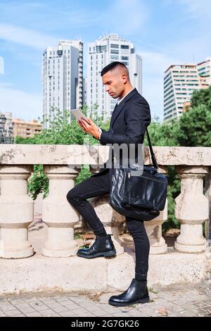 Side view of focused young Hispanic male executive manager in classy suit with bag reading information on tablet while standing near railing on urban Stock Photo