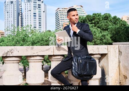 Side view of focused young Hispanic male executive manager in classy suit with bag reading information on tablet while standing near railing on urban Stock Photo
