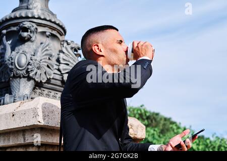 Low angle side view of young Hispanic male in formal black suit with mobile phone in hand enjoying refreshing takeaway drink while resting on urban st Stock Photo
