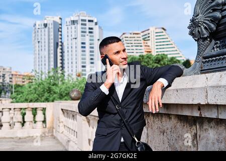 Confident young man in elegant formal suit talking on mobile phone and smiling while standing near sculpture on urban square Stock Photo