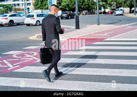 Full body of young well dressed male entrepreneur in classy suit and protective mask carrying briefcase and disposable cup of takeaway drink walking o Stock Photo