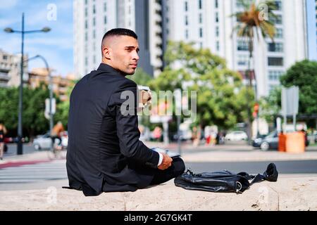 Low angle side view of serious pensive young Hispanic man in formal black suit looking away thoughtfully while sitting with digital gadget in hand on Stock Photo