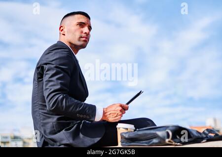 Low angle side view of serious pensive young Hispanic man in formal black suit looking away thoughtfully while sitting with digital gadget in hand on Stock Photo