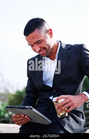Side view of happy young Hispanic man in elegant formal suit drinking takeaway beverage and browsing tablet while having free time and resting in park Stock Photo