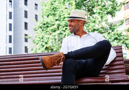 Confident handsome young Hispanic male in elegant clothes and hat sitting on bench and looking away while resting on city street Stock Photo