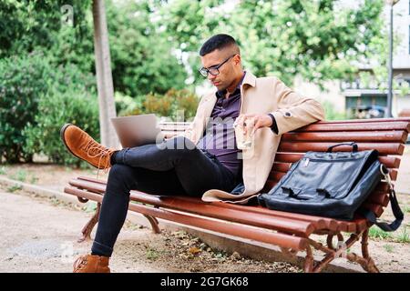 Focused young businessman in stylish outfit and glasses with cup of takeaway coffee working on laptop while sitting on bench in urban park Stock Photo